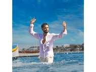 Homme en chemise blanche mouillée dans l'océan, bras levés, bateau et bâtiments en arrière-plan.