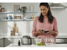 Charger l&#39;image dans la galerie, Femme dans la cuisine avec un robot culinaire, tenant un accessoire noir, souriante.
