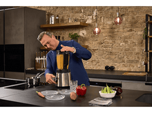 Charger l&#39;image dans la galerie, Un homme en chemise bleue mixe une soupe. Cuisine avec mur de briques et ingrédients sur le comptoir.
