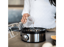 Charger l&#39;image dans la galerie, Une femme verse de l&#39;eau dans un cuiseur à œufs sur un comptoir, œufs visibles.
