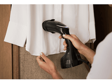 Charger l&#39;image dans la galerie, Une personne repasse une chemise blanche avec un défroisseur noir tenu dans la main droite.
