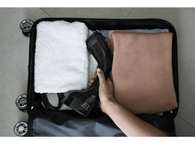 Charger l&#39;image dans la galerie, Une main tenant un défroisseur noir à l&#39;intérieur d&#39;une valise ouverte avec des vêtements pliés et une serviette.
