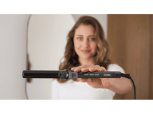 Charger l&#39;image dans la galerie, Une femme tient un fer à friser noir, souriant à la caméra. Un miroir est en arrière-plan.
