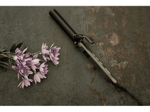 Charger l&#39;image dans la galerie, Un fer à friser noir à côté d&#39;un bouquet de fleurs violettes sur une surface sombre texturée.
