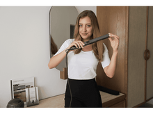 Charger l&#39;image dans la galerie, Une femme tenant un lisseur de cheveux devant un miroir dans une pièce.
