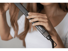 Charger l&#39;image dans la galerie, Une femme se lisse les cheveux avec un fer plat argenté. Le fer a un cordon noir.
