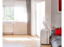 Charger l&#39;image dans la galerie, Un salon avec un climatiseur mobile, une fenêtre, un canapé et un sol en bois. Il y a de la lumière du soleil.
