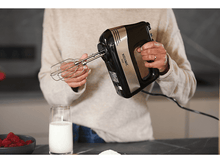 Charger l&#39;image dans la galerie, Une personne tenant un batteur à main noir et argenté. Un verre de lait et des framboises sur la table.
