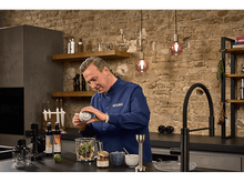 Charger l&#39;image dans la galerie, Chef en chemise bleue ajoutant du contenu à un mélangeur dans une cuisine. D&#39;autres ingrédients sont visibles.
