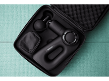 Charger l&#39;image dans la galerie, Pistolet de massage noir dans un étui, avec accessoires. Sur une surface verte texturée.
