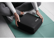 Charger l&#39;image dans la galerie, Une personne assise sur un tapis de yoga, tenant une mallette carrée noire avec le mot &#39;KOENIC&#39;.
