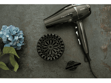 Charger l&#39;image dans la galerie, Sèche-cheveux noir avec accessoires, à côté d&#39;une fleur bleue sur une surface grise texturée.
