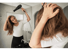 Charger l&#39;image dans la galerie, Une femme se sèche les cheveux devant un miroir. Elle porte une chemise blanche et une jupe noire. Cadre de salle de bain.
