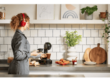 Charger l&#39;image dans la galerie, Femme utilisant une machine à café, portant un casque, avec de la nourriture et une décoration sur un comptoir de cuisine.
