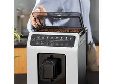 Charger l&#39;image dans la galerie, Une personne ouvrant le couvercle d&#39;une machine à café Krups, remplie de grains de café.
