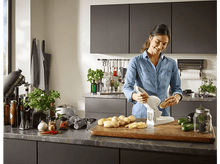 Charger l&#39;image dans la galerie, Femme tranchant des pommes de terre avec une trancheuse blanche dans une cuisine, légumes sur le comptoir.
