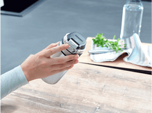 Charger l&#39;image dans la galerie, Une main tenant une bouteille d&#39;eau en métal. Le couvercle est noir avec un bouton blanc, la bouteille est argentée. Une table est dans la scène.
