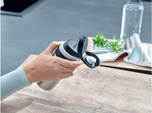 Charger l&#39;image dans la galerie, Main tenant une bouteille d&#39;eau noire et en acier inoxydable. Fond : table en bois, cuisine.
