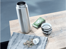 Charger l&#39;image dans la galerie, Une bouteille d&#39;eau en acier inoxydable, un sachet de thé et quelques feuilles de thé sur une table en bois.
