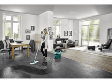 Charger l&#39;image dans la galerie, Une femme lave un sol en carrelage sombre dans un salon lumineux avec des meubles et des fenêtres.
