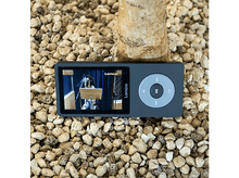 Charger l&#39;image dans la galerie, Un lecteur MP3 Lenco gris sur un lit de petits cailloux. L&#39;écran montre une personne en train de chanter.
