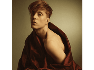 Un homme roux à l'épaule nue porte une robe de satin rouge, regardant la caméra.