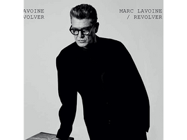 Image en noir et blanc de Marc Lavoine en manteau noir et lunettes, appuyé sur une surface.