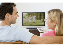 Charger l&#39;image dans la galerie, Couple regardant la télé. L&#39;homme et la femme sourient. La télé montre un couple marchant dans un champ vert.
