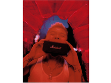 Charger l&#39;image dans la galerie, Une personne tenant un haut-parleur Marshall noir devant sa bouche, avec un fond abstrait rouge.
