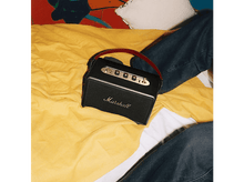 Charger l&#39;image dans la galerie, Un haut-parleur Marshall noir avec une poignée rouge est posé sur un lit jaune. Un jean et une couverture colorée en arrière-plan.
