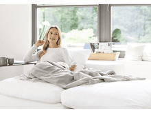 Charger l&#39;image dans la galerie, Femme sur un canapé blanc avec une couverture. Elle tient une télécommande. Une fenêtre est derrière elle.
