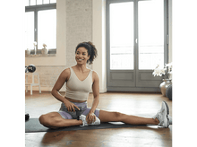 Charger l'image dans la galerie, Une femme assise sur un tapis utilise un pistolet de massage sur sa jambe, souriant dans une pièce lumineuse.
