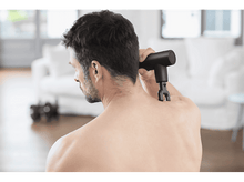 Charger l'image dans la galerie, Homme utilisant un pistolet de massage noir sur son épaule, à l'intérieur, avec canapé blanc.
