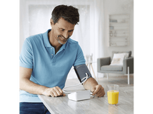 Charger l&#39;image dans la galerie, Homme vérifiant sa tension artérielle. Assis à une table avec du jus d&#39;orange. Appareil blanc. Fond clair.
