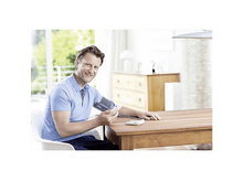 Charger l&#39;image dans la galerie, Homme assis à une table, prenant sa tension avec un appareil, souriant à la caméra.
