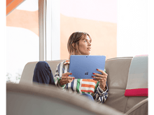 Charger l&#39;image dans la galerie, Une femme assise sur un canapé tient une tablette bleue, portant une chemise rayée. Elle regarde vers le haut, avec une fenêtre et un mur orange en arrière-plan.
