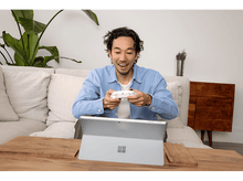 Charger l&#39;image dans la galerie, Un homme joue, tenant une manette blanche, regardant un ordinateur portable. Une plante et un canapé sont en arrière-plan.
