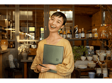 Charger l&#39;image dans la galerie, Une femme sourit, tenant un ordinateur portable et un stylo. Elle est dans un café, avec des étagères de bouteilles et de la déco.
