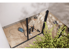 Charger l&#39;image dans la galerie, Une personne passant l&#39;aspirateur dans une pièce avec un aspirateur bleu. On peut voir beaucoup de plantes vertes.
