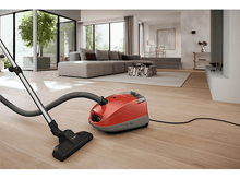 Charger l&#39;image dans la galerie, Un aspirateur orange dans une pièce avec des planchers en bois et un canapé, avec un tuyau d&#39;aspirateur.
