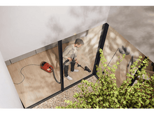 Charger l&#39;image dans la galerie, Une personne passe l&#39;aspirateur sur un sol en bois dans une pièce vitrée, entourée de plantes.
