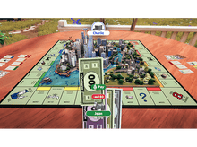Charger l&#39;image dans la galerie, Une partie de Monopoly en cours. Une joueuse nommée Jean paie Charlie. Le plateau a un modèle de ville au centre.
