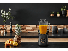 Charger l&#39;image dans la galerie, Mixeur avec jus d&#39;orange sur un plan de travail noir, avec fruits et plantes.

