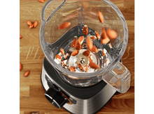 Charger l&#39;image dans la galerie, Un mixeur avec des amandes qui y tombent. Le mixeur est sur une surface en bois.
