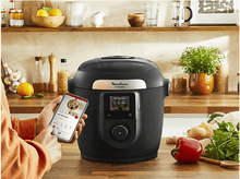 Charger l&#39;image dans la galerie, Personne utilisant un smartphone pour contrôler un multicuiseur intelligent Moulinex Cookeo noir sur bois.
