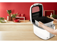 Charger l&#39;image dans la galerie, Une personne retirant un récipient noir d&#39;un appareil blanc, avec un arrière-plan de cuisine rouge.
