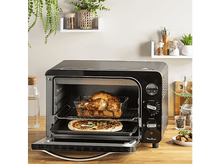 Charger l&#39;image dans la galerie, Un four Moulinex noir avec poulet rôti et pizza. Étagères, bocaux et table en bois en arrière-plan.
