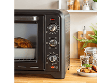 Charger l&#39;image dans la galerie, Four noir avec de la nourriture à l&#39;intérieur. Boutons sur le devant. Surface en bois, fond blanc.
