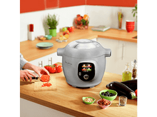 Charger l&#39;image dans la galerie, Une personne coupe une tomate à côté d&#39;un Moulinex Cookeo gris. Légumes et bols sur la table.
