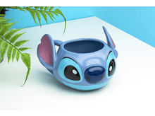 Charger l&#39;image dans la galerie, Un mug Stitch bleu avec de grandes oreilles et des yeux. Il est sur une surface blanche avec des plantes vertes.
